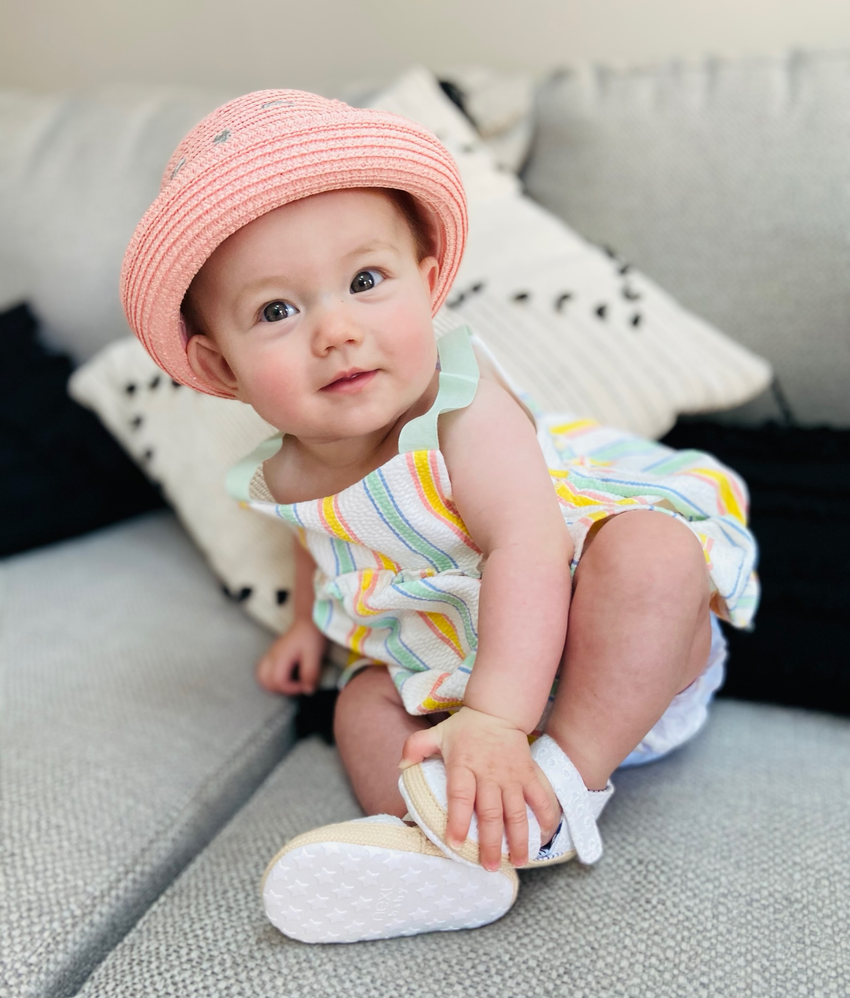 Orla sitting on the sofa in a pink sun hat and striped outfit, holding her shoe